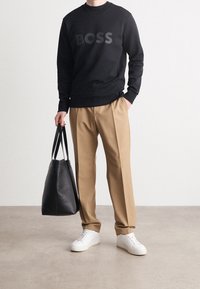 Felpa nera con logo "BOSS", pantaloni beige a pieghe, sneakers bianche, che tiene una borsa a tracolla in pelle nera. Texture lisce e design casual.