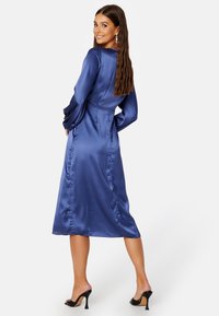 Sukienka midi z satyny w niebieskim kolorze z długimi, rozkloszowanymi rękawami oraz dopasowaną talią. Gładka tekstura, długość do kolan, noszona z czarnymi sandałami na obcasie.