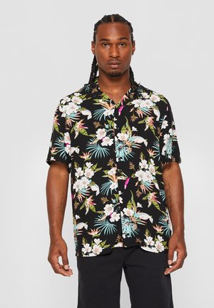 Man with braided hair wearing a black short-sleeve button-up shirt with colorful tropical floral pattern and black pants.