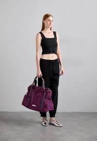 Kvinne i svart crop top og joggebukser som holder en stor lilla treningsbag, står mot en enkel grå vegg og betonggulv.