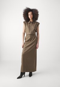 Camicia verde oliva con bottoni e maniche corte abbinata a una lunga gonna coordinata. Il modello indossa stivali neri con tacco. Tessuto liscio.
