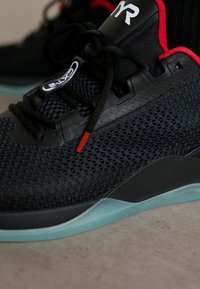 Czarne siatkowe buty sportowe z czerwonymi akcentami, czarne sznurowadła z czerwonymi końcówkami, białe logo "TYR" na języku i jasnoniebieska podeszwa na szarym podłożu.