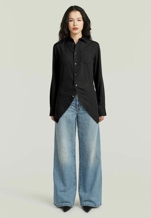 Frau trägt ein langes schwarzes Hemd mit Knöpfen, asymmetrischem Saum und weitem, ausgewaschenem blauen Jeansstoff und steht vor einem schlichten Hintergrund.