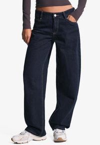 Tmavě modré široké džíny z denimu, s klasickým designem pěti kapes a kontrastním šitím. Nosené se světlými sportovními botami.