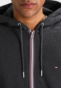 Hoodie zippé gris avec une fermeture éclair contrastante rouge et blanche. Dispose de cordons de serrage, d'une texture douce et d'un petit logo sur la poitrine.