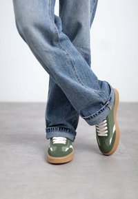 Zapatillas verdes con acentos blancos y suela de goma marrón, combinadas con jeans de denim azul sueltos, que presentan un dobladillo deshilachado y un corte relajado.