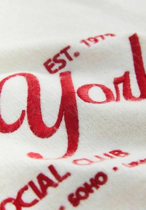 Valge kangas punase tikandiga tekstiga "May York" ja "EST. 1973 SOCIAL CLUB" erinevates kirjatüüpides. Märgatav peen tekstuur.