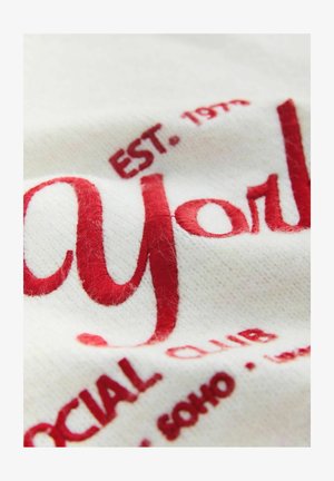 Valge kangas punase tikandiga tekstiga "May York" ja "EST. 1973 SOCIAL CLUB" erinevates kirjatüüpides. Märgatav peen tekstuur.