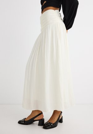 Žena koja nosi bijelu maksi suknju visokog struka s naborima i crne slingback štikle sa srebrnim zakovicama, na jednostavnoj bijeloj pozadini.