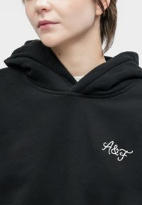 Inimene mustas kapuutsiga, millel on rinnas valge "A&F" tikand, näidates alanägu ja kaela ühetoonilise tausta ees.