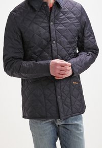 Mann trägt eine dunkle gesteppte Barbour-Jacke mit Druckknöpfen und Jeans, die Hände auf der Hüfte verschränkt, steht vor einem schlichten Hintergrund.