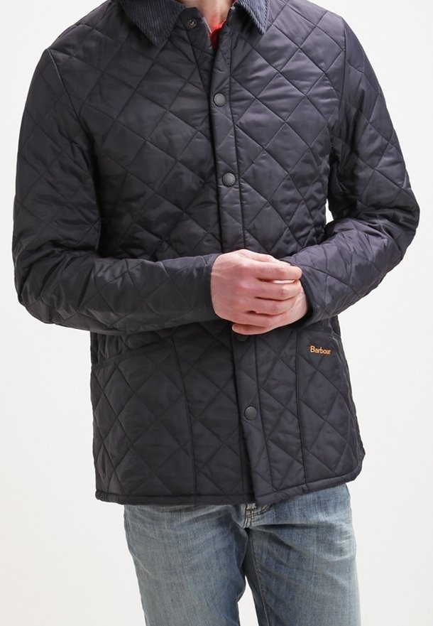 Mann trägt eine dunkle gesteppte Barbour-Jacke mit Druckknöpfen und Jeans, die Hände auf der Hüfte verschränkt, steht vor einem schlichten Hintergrund.