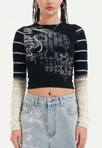 Maglietta nera a maniche lunghe con grafica di un teschio, accenti floreali e maniche a strisce sfumate da chiaro a scuro. Abbinata a jeans di denim blu chiaro.