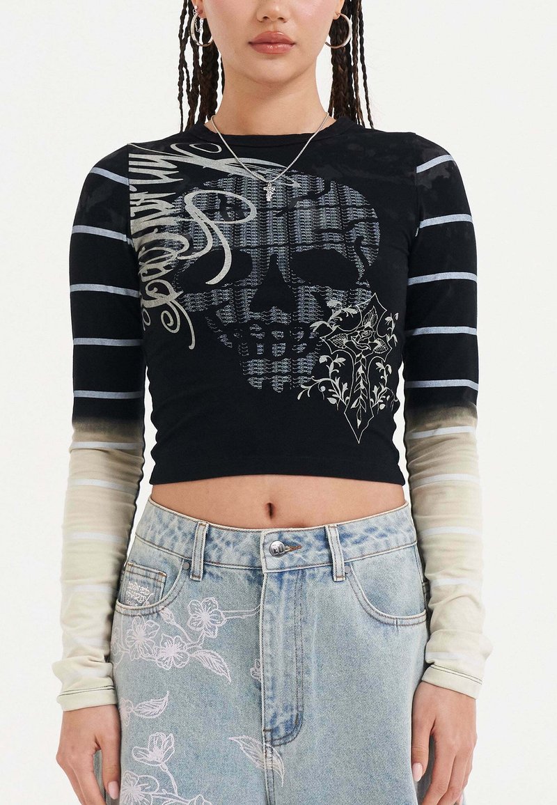 Maglietta nera a maniche lunghe con grafica di un teschio, accenti floreali e maniche a strisce sfumate da chiaro a scuro. Abbinata a jeans di denim blu chiaro.