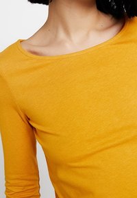 Dlouhý rukáv v jasně žluté tkanině, s širokým výstřihem a hladkou texturou bez viditelných vzorů nebo akcentů.