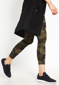 Green camo leggings with a fitted design, made from stretchy fabric. Worn with a black tunic and black sneakers with three straps.