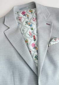 Hellgrauer maßgeschneiderter Blazer aus strukturiertem Stoff. Innen ist er mit einem Blumenmuster gefüttert und enthält ein passendes Einstecktuch. Subtile lila Nähte setzen Akzente.