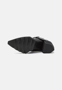 Zapato de tacón alto negro con una suela de goma texturizada, que presenta una puntera ancha y un tacón angular, acentuado por una hebilla simple en la correa.
