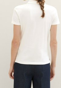 Vit kortärmad polo med krage, åtsittande design, strukturerat bomullsmaterial och rak nederdel, tillsammans med mörka denimbyxor.