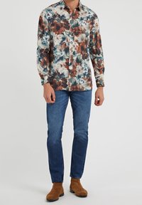 Chemise à manches longues avec boutons, à motif floral dans des teintes de marron, vert et blanc ; associée à un jean bleu et des bottines brunes.