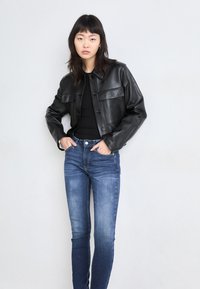 Μαύρο δερμάτινο κοντό μπουφάν με κλείσιμο με κουμπί, τσέπες στο στήθος, σε συνδυασμό με εφαρμοστά μπλε τζιν. Απλή φόντο.