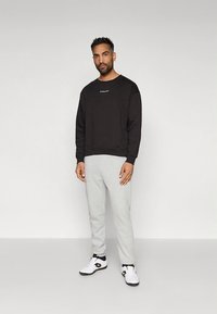 Μαύρο φούτερ με στρογγυλή λαιμόκοψη και μικρό λευκό λογότυπο, σε συνδυασμό με ανοιχτό γκρι φόρμες και λευκά αθλητικά παπούτσια. Απλός σχεδιασμός.