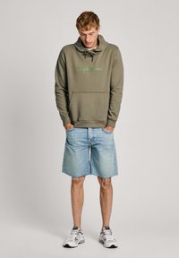 Sudadera color verde oliva con un bolsillo frontal y capucha con cordón, con la inscripción "Pepe Jeans" en letras verde claro. Combinada con pantalones cortos de mezclilla azul claro.