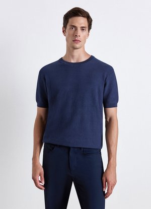Giovane uomo che indossa una maglia a maniche corte lavorata a maglia di colore blu scuro con trama e pantaloni scuri, in piedi davanti a uno sfondo chiaro e uniforme.