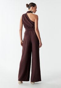 Combinaison bordeaux avec un design à une épaule, tissu texturé, coupe évasée et détail de col tressé, portée avec des sandales à talons.