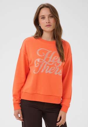 Femme aux longs cheveux bruns portant un sweat-shirt orange avec le texte "Hey There" et un pantalon noir, debout devant un fond clair uni.