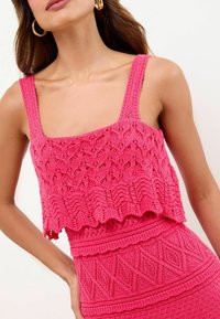 Roze gebreide crop top met een vierkante halslijn, gekartelde onderrand en ingewikkelde opengewerkte patronen. Textuurbandjes maken het ontwerp compleet.