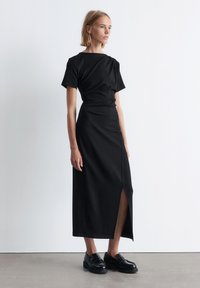 Schwarzes Midi-Kleid mit kurzen Ärmeln, das einen drapierten Oberteil, einen Schlitz an der Seite und eine figurbetonte Silhouette aufweist. Getragen mit schwarzen Loafers.