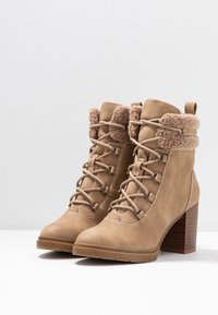 Beżowe zamszowe botki za kostkę na grubych obcasach, z sznurowanym przodem i wełnianym wykończeniem na górze, ustawione obok siebie na białej powierzchni.