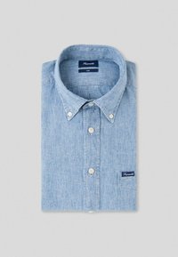 Chemise bleu clair texturée à manches longues avec boutons, col boutonné et petite poche montrant l’étiquette de la marque pliée sur fond gris.