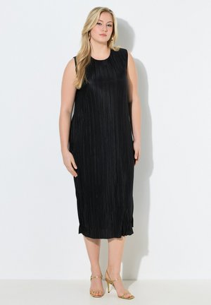 Blondynka w czarnej plisowanej sukience midi bez rękawów i złotych sandałach na wysokim obcasie, stojąca przy białej ścianie.