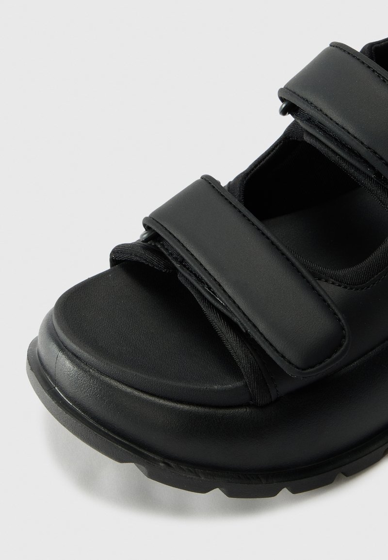 Schwarze Sandale mit zwei breiten Klettverschlussriemen, einer strukturierten Sohle und einer abgerundeten Zehenform. Aus synthetischem Material mit matter Oberfläche gefertigt.