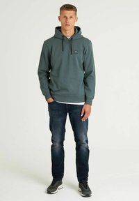 Felpa con cappuccio di colore verde scuro, realizzata in morbido misto cotone, con tasca frontale e un piccolo logo sul petto; abbinata a jeans scuri.