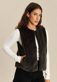 Vrouw met lang haar, gekleed in een zwart nepbont vest over een witte langwerpige top en zwarte broek, staande met één hand in de zak.