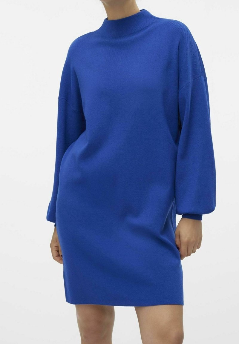 Femme portant une robe-pull à manches longues bleu roi avec un col montant et une coupe décontractée, debout devant un fond blanc uni.