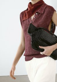 Viininpunainen neulepusero ilman hihoja, jossa on kaulus ja napit, yhdistetty valkoisiin teksturoituihin housuihin ja mustaan kudottuun clutch-laukkuun. Punainen huivi korostaa asukokonaisuutta.