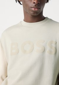 Sudadera beige con un logotipo "BOSS" texturizado y en relieve de un color a juego. Diseño simple de cuello redondo, hecho de una tela suave y lisa.