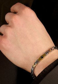 Bracciale in tonalità argento e oro con anelli intrecciati e un pannello centrale testurizzato con un logo nero, indossato su una mano.