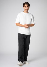 Weißes T-Shirt mit kurzen Ärmeln und Rundhalsausschnitt, kombiniert mit dunkler Jeanshose und weißen Sneakern. Das Modell steht mit gefalteten Händen vor einem neutralen Hintergrund.