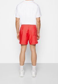 Muž v jasně červených plavkách s kapsou na zadní straně, bílých ponožkách do poloviny lýtek, bílých teniskách a jednoduchém bílém tričku, stojící na jednobarevném pozadí.