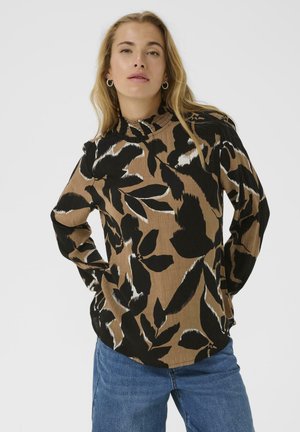 Woman posing with hands on hips, wearing hoop earrings, a brown and black leaf-patterned long-sleeve top, and blue jeans against a white background.