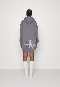 Grå överdimensionerad hoodie-klänning med en rygglogotyp "Calvin Klein Jeans." Har huva, lösa ärmar och sidofack. Paret med vita knähöga stövlar.
