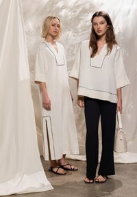 Deux mannequins portent des vêtements crème clair avec des bordures noires : l'un porte une robe mi-longue fendue, l'autre un haut à manches courtes sur un pantalon noir.