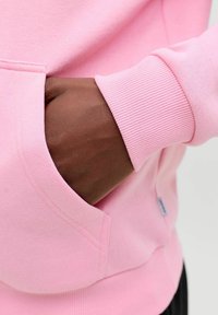 Felpa rosa con polsini a coste, caratterizzata da una tasca frontale. Primo piano che mostra la texture liscia e i dettagli delle cuciture.