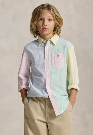 Enfant blond avec des cheveux mi-longs portant une chemise rayée multicolore boutonnée et un pantalon beige, mains dans les poches, fond neutre.