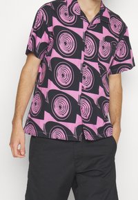 Lyhythihainen paita mustana ja vaaleanpunaisena, jossa pyörteisiä kuvioita, napitettava etuosa, rintatasku ja rento istuvuus. Puuvillakankaan tekstuuri.